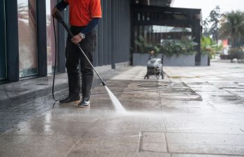 A specialist pressure washing a sidewalk in front of a commercial building.